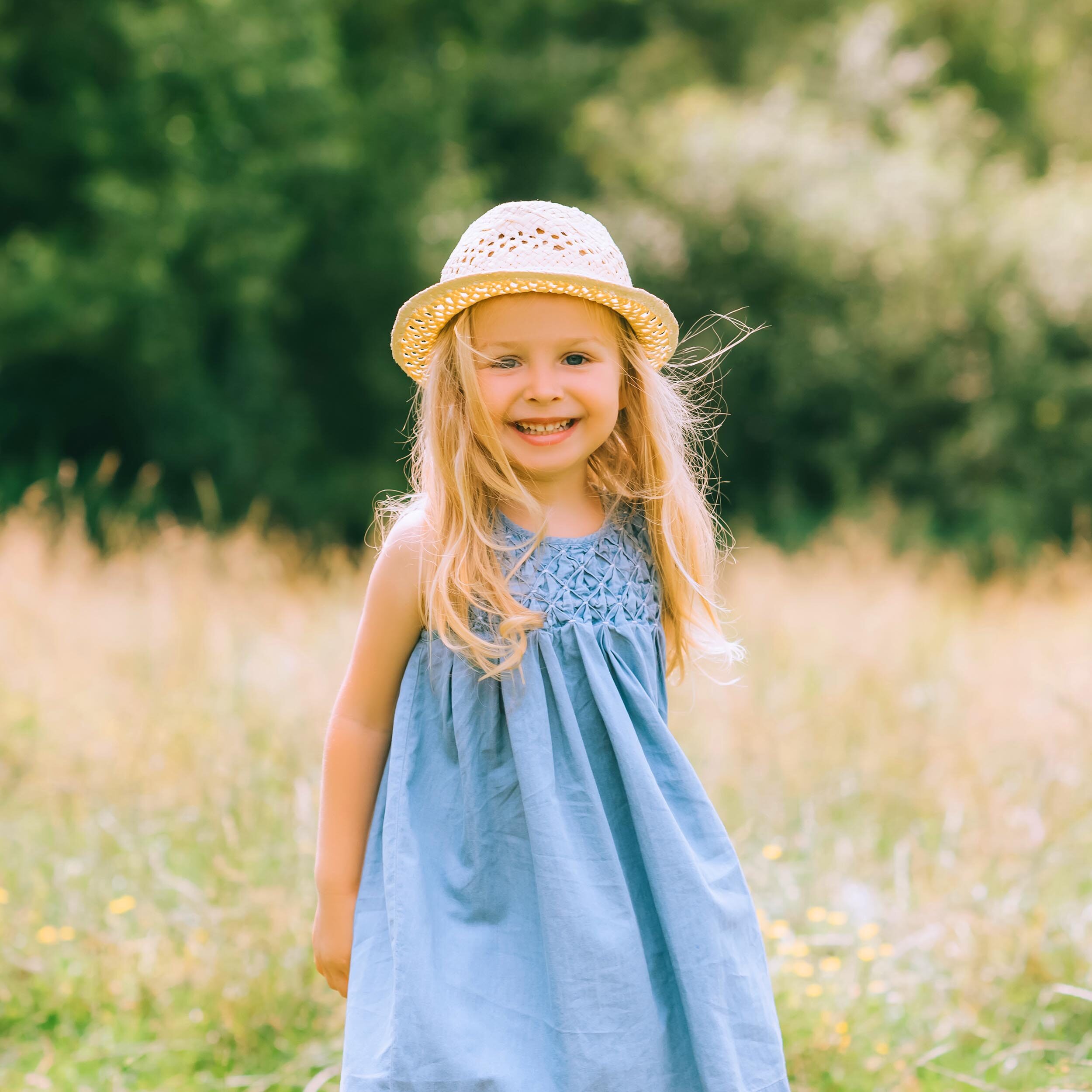 adorable blonde smiling child in dress and straw hat in field